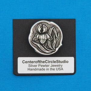 Könnte beinhalten: Eine Brosche aus Silberzinn in Form einer Iris-Blume, handgefertigt in den USA. Die Brosche ist rund und zeigt ein detailliertes Design einer Iris-Blume mit Blättern. Die Brosche befindet sich auf einem schwarzen Hintergrund mit einem weißen Etikett, auf dem "CenteroftheCircleStudio Silver Pewter Jewelry Handmade in the USA" steht.