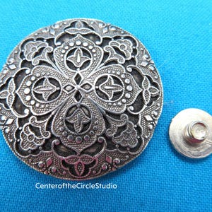 May include: Silver-toned metal button with a decorative floral design. The button has a round shape and a raised center. The button is attached to a silver-toned metal post.