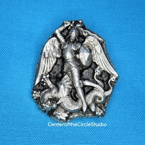 May include: Silver tone brooch depicting an angel slaying a dragon. The angel is wearing armour and has wings. The dragon is curled around the angel's legs. The brooch has a black enamel background.