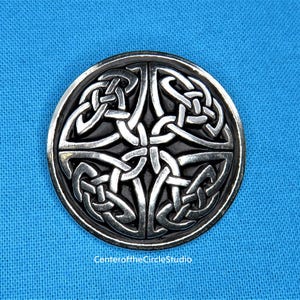 May include: A silver Celtic knot design brooch with a black background. The knot design is intricate and detailed.