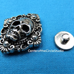 May include: Silver skull charm with ornate details, set in a square frame. The charm is attached to a silver screw back.
