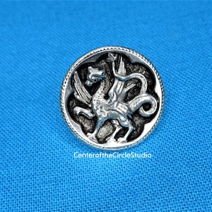 A silver metal pin with a detailed engraved design of a winged creature, possibly a dragon or mythical beast, on a blue fabric background.