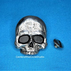 May include: A silver skull with black eyes, a decorative button or embellishment for clothing or crafts.