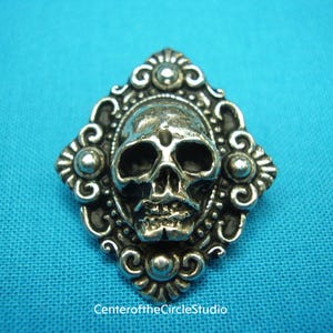 Könnte beinhalten: Ein silberner Totenkopf-Anhänger mit einem verzierten, rautenförmigen Rahmen. Der Totenkopf ist detailliert mit Augenhöhlen, einer Nase und einem Mund.