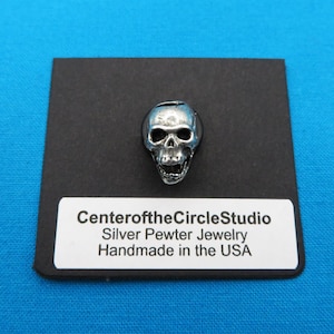 May include: A silver pewter skull charm pendant, handmade in the USA. The skull is detailed with a textured surface and has a realistic appearance. The charm is displayed on a black background with the text "CenteroftheCircleStudio Silver Pewter Jewelry Handmade in the USA".