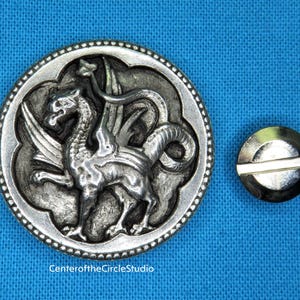 May include: A silver-toned metal button with a detailed design of a winged dragon. The button has a textured, ornate border.