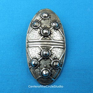 May include: A silver oval brooch with a textured surface and raised circular accents. The brooch has a geometric design with a central cross.