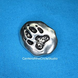 May include: A silver charm with a detailed paw print design. The charm is shaped like a rounded stone and has a textured surface.