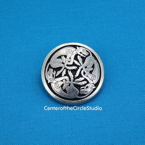 May include: A round, silver-colored metal button with a Celtic knot design. The button features intertwined animal figures and intricate knotwork. The text "Center of the Circle Studio" is at the bottom of the image. The button is set against a blue background.