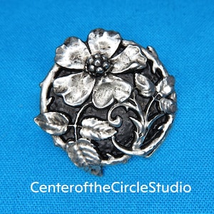 May include: A silver-toned metal brooch featuring a detailed floral design. The flower is a stylized rose with a black background. The brooch is a circular shape.