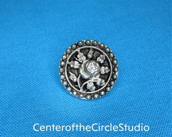 May include: A silver button with a raised design of a rose and leaves. The button has a beaded edge.
