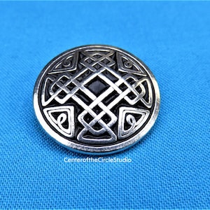 Peut inclure: Un bouton rond argenté avec un motif de nœud celtique. Le nœud est de couleur noire et est centré sur le bouton.