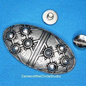 May include: A silver oval-shaped metal buckle with a geometric design and raised dots. The buckle is accompanied by two silver metal fasteners.
