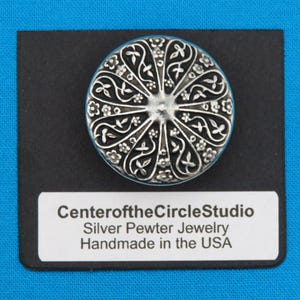 May include: A silver pewter jewelry piece with an intricate floral design. The jewelry is handmade in the USA. The text "Center of the Circle Studio" is printed on a white label.