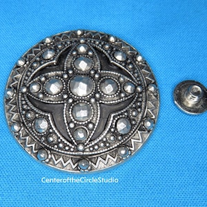 May include: A silver metal button with a black center and a raised design of a four-pointed star. The button has a decorative border and a small silver metal post on the back.