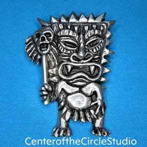 May include: Silver tiki brooch with a fierce expression, holding a skull in one hand. The brooch has a detailed design with sharp edges and a textured surface.