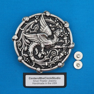 May include: A silver pewter belt buckle with a detailed design of a griffin with wings spread, surrounded by intricate floral patterns. The buckle is round and has a black background. The text "CenteroftheCircleStudio Silver Pewter Jewelry Handmade in the USA" is printed on a black rectangle below the buckle.