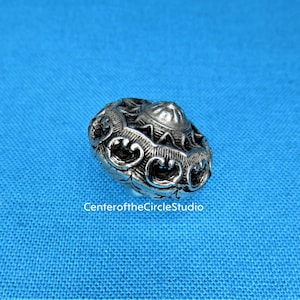 May include: A silver metal button with an intricate, ornate design. The button has a raised centre and a circular shape.