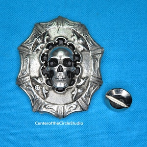 May include: A silver-toned metal skull with ornate detailing, set within a circular frame with a decorative design. The skull has black eyes and a detailed bone structure. The frame has a textured surface and a raised edge. A small silver-toned screw is included.