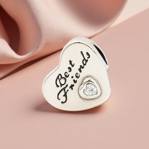 K&ouml;nnte beinhalten: Ein silberner Charm in Herzform mit dem Schriftzug "Best Friends" in Schreibschrift. In der Mitte befindet sich ein kleineres Herz mit einem klaren Stein. Der Charm ist f&uuml;r ein Armband oder eine Halskette gedacht.