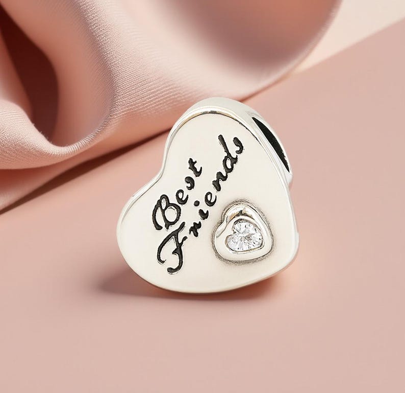 K&ouml;nnte beinhalten: Ein silberner Charm in Herzform mit dem Schriftzug "Best Friends" in Schreibschrift. In der Mitte befindet sich ein kleineres Herz mit einem klaren Stein. Der Charm ist f&uuml;r ein Armband oder eine Halskette gedacht.