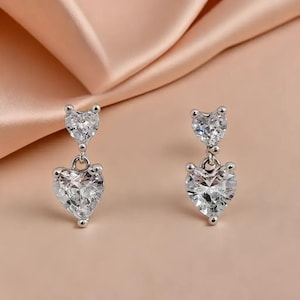 May include: A pair of silver drop earrings, each with two clear, heart-shaped cubic zirconia stones. The larger heart hangs below a smaller one. The earrings are set against a soft, peach-coloured background, creating a delicate and romantic aesthetic.