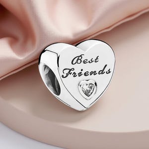 K&ouml;nnte beinhalten: Ein silberner herzf&ouml;rmiger Charm-Anh&auml;nger mit dem eingravierten Schriftzug "Best Friends" auf der Vorderseite. Ein kleines, klares Kristallherz befindet sich unter dem Text. Der Charm wird auf einer rosa Oberfl&auml;che pr&auml;sentiert.