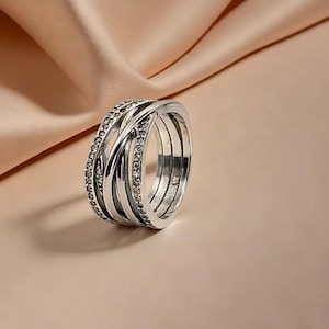 May include: A silver ring with an interwoven design. The ring has multiple bands, some set with small, clear gemstones. The ring is displayed against a soft, peach-coloured background, highlighting its intricate details and reflective surface.