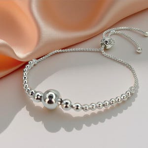 May include: A silver bracelet with a design of small, round beads and a larger central bead. The bracelet has an adjustable sliding closure with two small, round end beads. The jewellery is shown against a soft, peach-coloured background.
