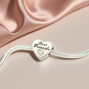 K&ouml;nnte beinhalten: Ein silberner Charm-Anh&auml;nger mit herzf&ouml;rmigem Anh&auml;nger. Der Anh&auml;nger tr&auml;gt die Aufschrift "Best Friends" und ein kleines Herz mit einem klaren Stein. Der Charm befindet sich an einem silbernen Armband, vor einem rosa Satinhintergrund.