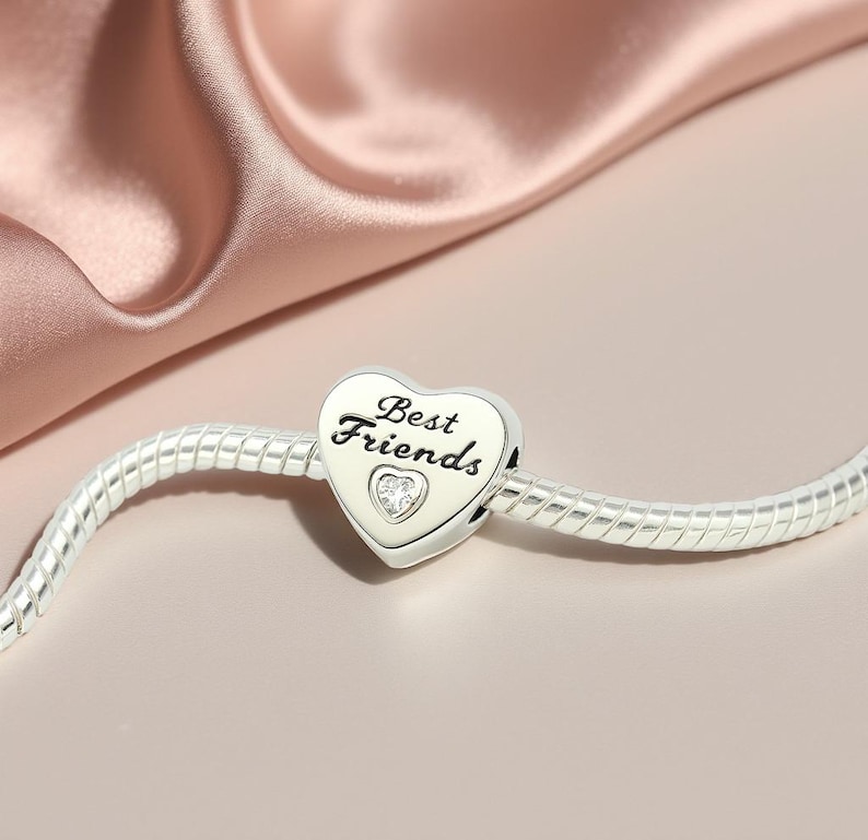 K&ouml;nnte beinhalten: Ein silberner Charm-Anh&auml;nger mit herzf&ouml;rmigem Anh&auml;nger. Der Anh&auml;nger tr&auml;gt die Aufschrift "Best Friends" und ein kleines Herz mit einem klaren Stein. Der Charm befindet sich an einem silbernen Armband, vor einem rosa Satinhintergrund.