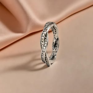 Puede incluir: Un anillo plateado con un diseño trenzado. El anillo está adornado con pequeñas piedras preciosas transparentes. El anillo se encuentra sobre un fondo suave de color melocotón, resaltando sus intrincados detalles y su estilo elegante.