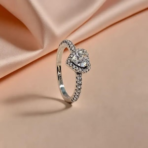 May include: A silver ring featuring a heart-shaped central stone, encircled by smaller stones. The band is also embellished with tiny stones. The ring is displayed against a soft, pink backdrop.