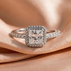 May include: A silver-toned ring featuring a square-cut centre stone surrounded by a halo of small round stones. The band is also set with a row of small round stones along each side, creating a continuous sparkling appearance on the metal band.