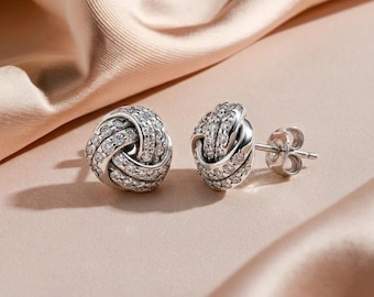 May include: A pair of silver-toned stud earrings featuring a knotted design. Each earring is encrusted with numerous small, clear, round-cut stones that cover the entire surface of the knot, and includes a standard metal post and butterfly back.