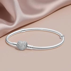 May include: A silver bracelet featuring a heart-shaped charm, encrusted with small, sparkling stones. The bracelet has a snake chain design. It is presented on a pink surface.