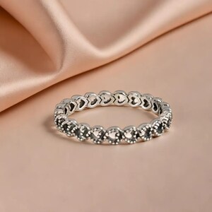 Könnte beinhalten: Ein silberner Ring mit einem sich wiederholenden Herzmuster. Der Ring ist vor einem weichen, rosa Hintergrund mit einem seidigen Stoff im Hintergrund platziert. Das Design des Rings besteht aus einer Reihe kleiner Herzen.