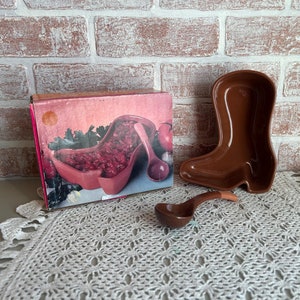May include: A brown ceramic boot-shaped serving dish with a matching brown ceramic ladle. The dish is filled with red fruit salad and is sitting on a white crocheted doily. The dish is in its original box, which is pink and has a picture of the dish on it.