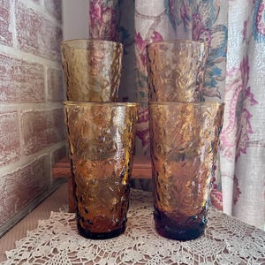 May include: A set of four amber-colored glass tumblers featuring a textured, pebbled surface. The glasses are arranged in two rows, with two shorter tumblers in the back and two taller, cylindrical tumblers in the front, all made of translucent glass.