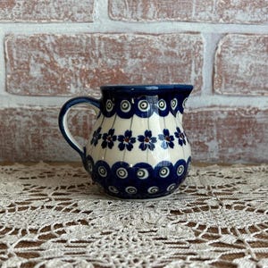 May include: A small, ceramic pitcher with a handle, featuring a blue and white floral pattern. The pitcher has a rounded body and a slightly flared rim. The design includes blue flowers and circular accents. The pitcher is approximately 4 inches tall.