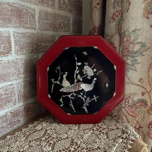 May include: An octagonal box featuring a vibrant red frame and a black central panel. The black panel is decorated with an intricate inlay of white and iridescent shell depicting two birds perched on flowering branches with delicate blossoms.