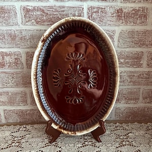 May include: An oval, brown ceramic serving platter with a decorative floral design in the center. The platter has a textured rim and is displayed on a brown stand. The background features a brick wall.