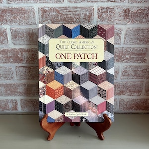 May include: A book titled "The Classic American Quilt Collection: One Patch" displayed on a wooden stand. The book cover features a colorful patchwork design in various colors and patterns. The background is a brick wall.