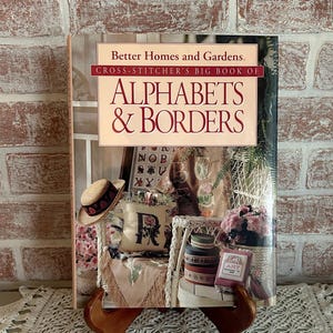 May include: A hardcover book titled "Better Homes and Gardens Cross-Stitcher's Big Book of Alphabets & Borders" is displayed on a wooden stand. The cover features a collage of cross-stitch designs and a straw hat. The book is open, revealing more cross-stitch patterns.