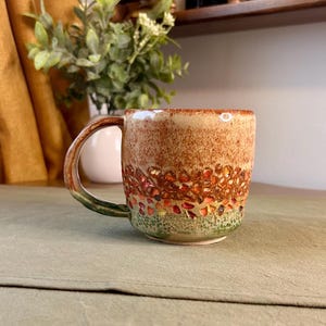 May include: A ceramic mug featuring a speckled beige upper section, a middle band of textured orange and brown leaf-like shapes, and a green speckled base. The handle is curved and colored in shades of brown and green, matching the mug's base.