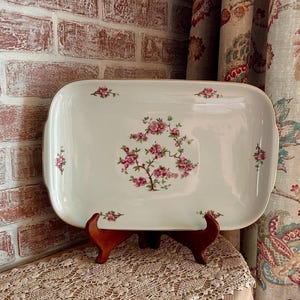 May include: A rectangular white porcelain serving platter featuring a central floral motif of pink blossoms on brown branches. Smaller matching floral sprigs are positioned at the four corners and the center of each side, finished with a gold rim.