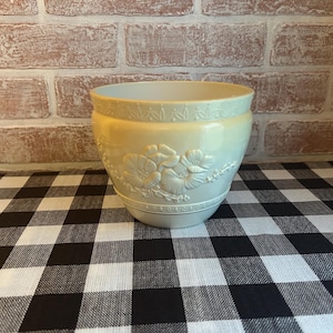 May include: A white ceramic planter with a raised floral design. The planter has a smooth, glossy finish and is sitting on a black and white checkered surface.