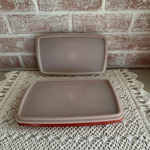 May include: Two rectangular Tupperware containers with lids. The lids are a light mauve color with a red rim on the bottom container. The containers are on a white lace doily in front of a brick wall.