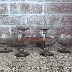 May include: A set of six clear glass coupes with wide, shallow bowls and short stems featuring a spherical accent above a circular base. The glassware is arranged in two rows, with two glasses in the back and four glasses in the front, showcasing a smooth, transparent finish throughout each piece.