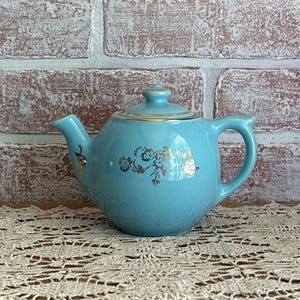 May include: A vintage, light blue teapot with a matching lid. The teapot features gold floral accents and a gold rim. The teapot is sitting on a lace doily in front of a brick wall.